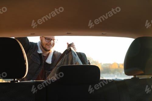 Preview: a smiling man taking things out of his car’s trunk, as viewed from within the car