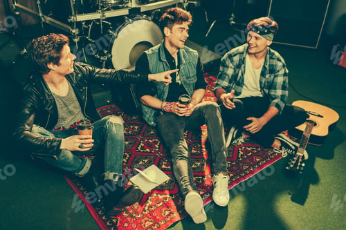 Preview: overhead view of cheerful rock band holding paper cups and talking while sitting on carpet