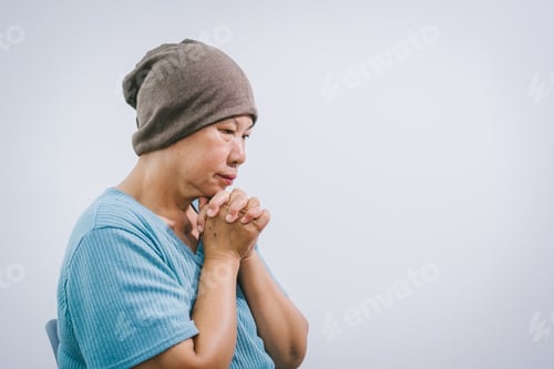 Preview: An elderly Asian woman wearing a gray-brown beanie, a light blue shirt,and dark pants sits quietly