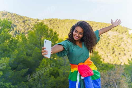 Preview: Curly haired woman smiling, taking a selfie with phone and feeling the freedom with the LGBT flag
