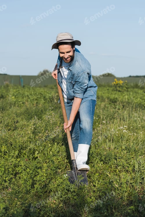 Preview: full length view of pleased farmer in brim hat and denim clothes digging in field