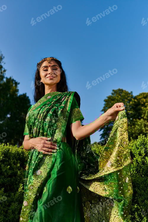Preview: cheerful and modern young indian woman touching sari with pattern near green plants in park outdoors