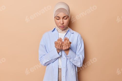 Preview: Calm religious Muslim woman makes traditional prayer to God keeps hands opened wears hijab and blue