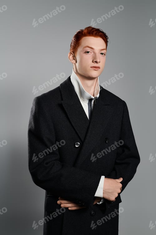 Preview: Young man with bright red hair poses confidently in a winter coat against grey