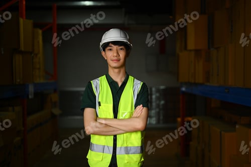 Preview: Confident male warehouse worker standing with arms crossed aisle between rows.