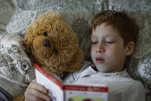 Preview: Child 6-7 years old red-haired boy lies in bed reading a book before going to bed with a teddy bear