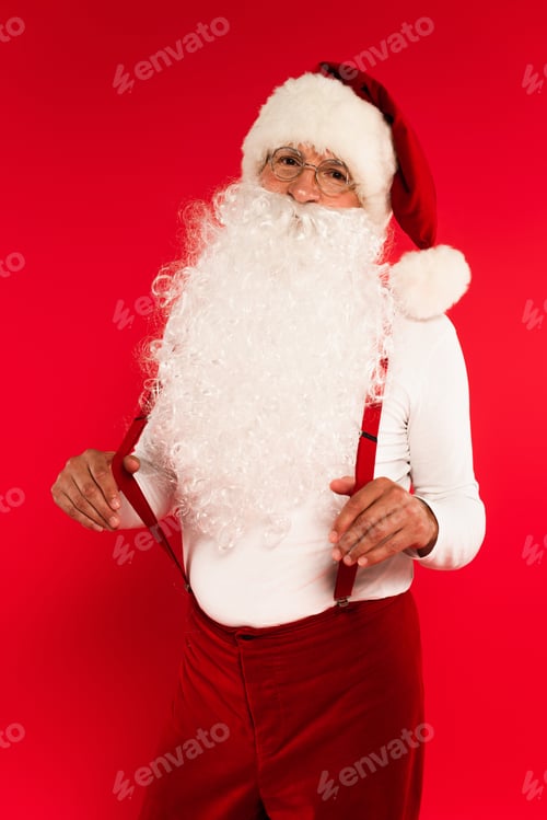 Preview: Bearded santa claus in eyeglasses holding suspenders and looking at camera on red background
