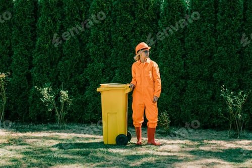 Preview: Stylish scavenger woman in orange suit, hardhat, and sunglasses with yellow can in outdoor