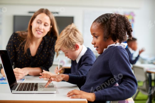 Preview: A female school teacher helping a boy and girl using a laptop computer in primary school class