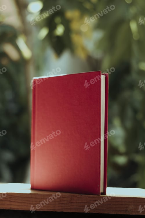 Preview: Vertical shot of a book with a red cover on a wooden fence in a garden under the sunlight