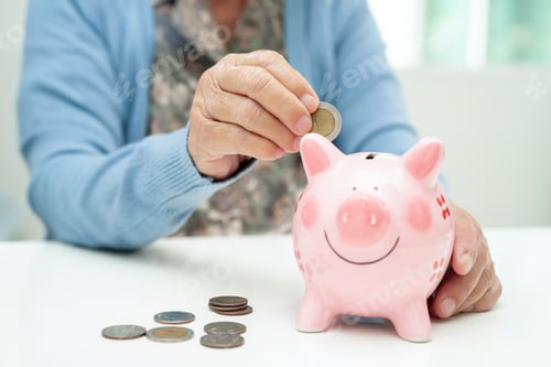 Preview: Asian elderly woman putting coin into pink piggy bank for saving money and insurance, poverty.