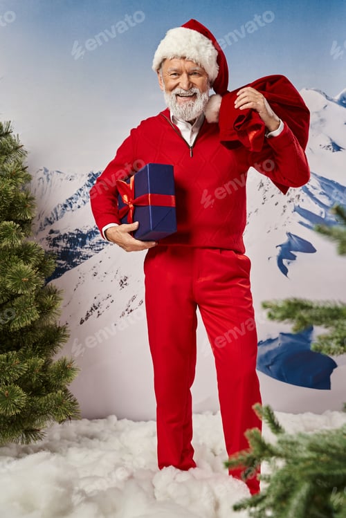 Preview: happy modern Santa with white beard in red outfit posing with gift and present bag, winter concept