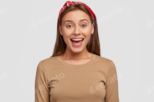 Preview: Happy green eyed European woman with pleased expression, keeps mouth opened, wears red bandana, beig