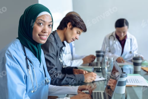 Preview: Mixed race female surgeon in hijab looking at camera while using laptop at table in hospital.