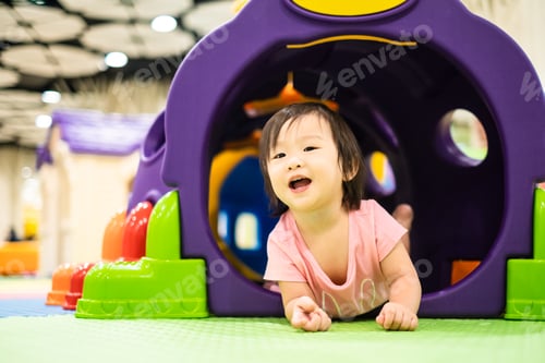 Preview: Little cute Asian toddler girl crawling out from colorful toy tunnel at playground.