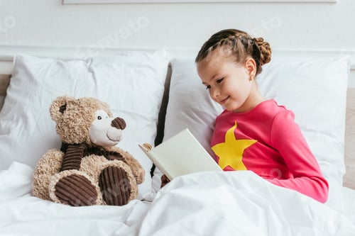Preview: adorable child reading book to teddy bear while sitting on bed