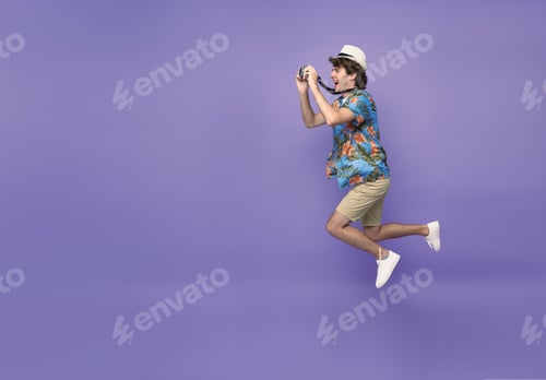 Preview: Young man tourist smiling and jumping with camera in hand isolated on purple background.