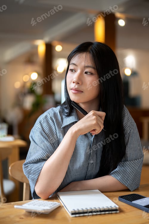 Preview: Asian woman holding pen under chin looking around with calculator and notebook on wood table in cafe
