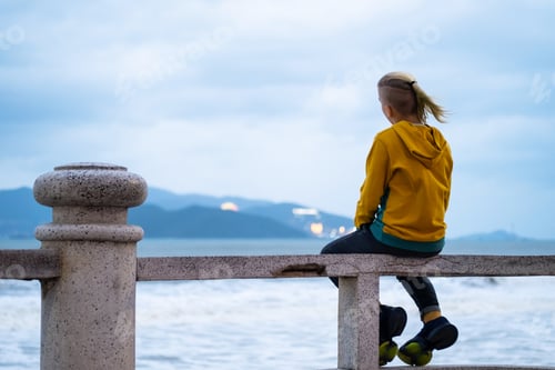 Preview: Lonely teenager looking to sea sitting on the waterfront. Rear view of teenager against seascape.