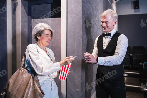 Preview: Elegant usher checking ticket of woman holding popcorn bucket in movie theater