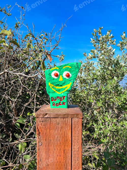 Preview: Green painted Gumby rock on wood stake in bushes under blue sky.