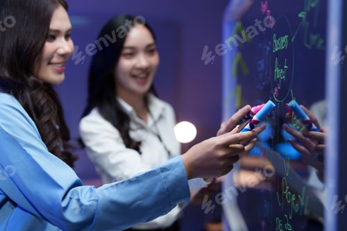 Preview: Businesswomen planning strategy, writing on transparent whiteboard in office at night