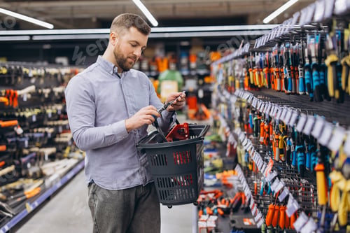 Preview: Man holding a screwdriver and a shopping basket, browsing through rows of various hand tools