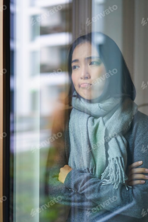Preview: Portrait of sad ill korean woman looking out of the window, staying at home due to coronavirus