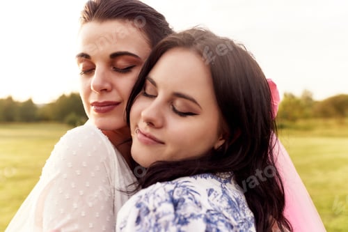 Preview: Charming young bride in a white dress with white veil and a bridesmaid with pink veil hug gently