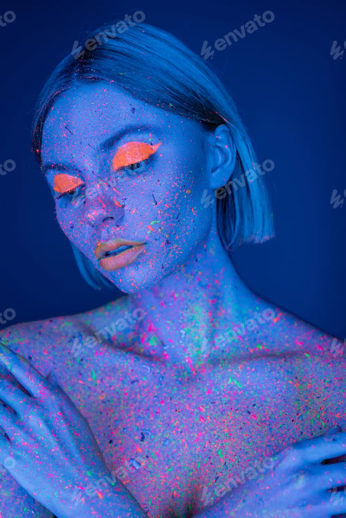 Preview: nude woman in bright neon makeup and fluorescent paint splatters posing isolated on dark blue