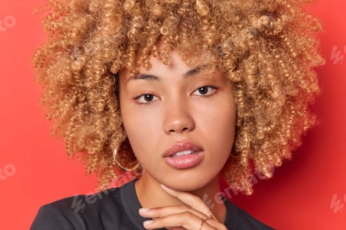 Preview: Close up portrait of serious curly haired woman touches jawline has healthy soft skin wears no makeu