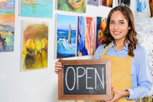 Preview: happy female artist holding signboard with word open in workshop
