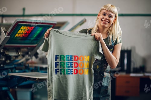 Preview: Happy print shop worker showing finished t-shirt at the camera,