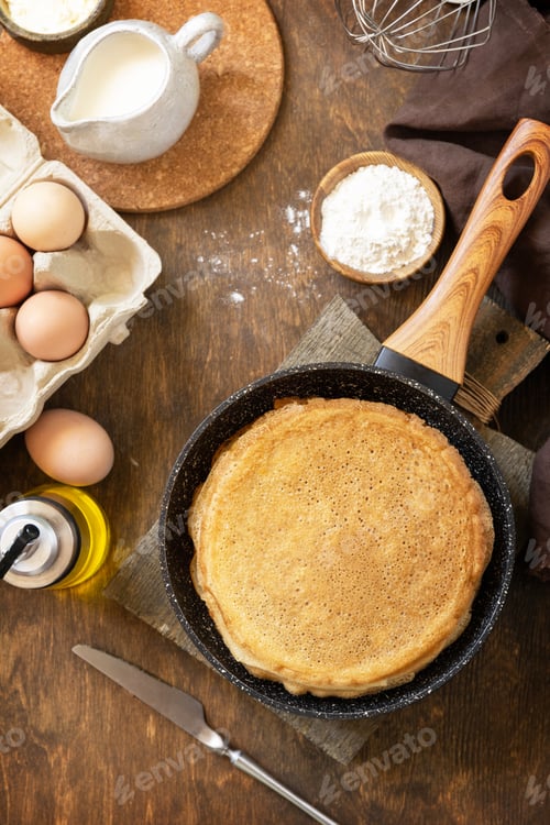 Preview: Delicious homemade crepes or pancakes in a frying pan and ingredients on a rustic table.