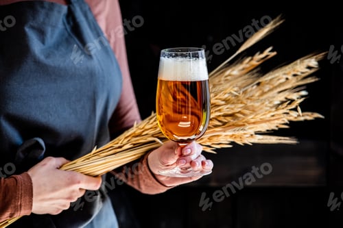 Preview: brewer in apron holds glass of beer and wheat spikelets