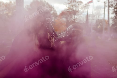 Preview: Couple in Halloween costumes and masks in a graveyard behind pink smoke