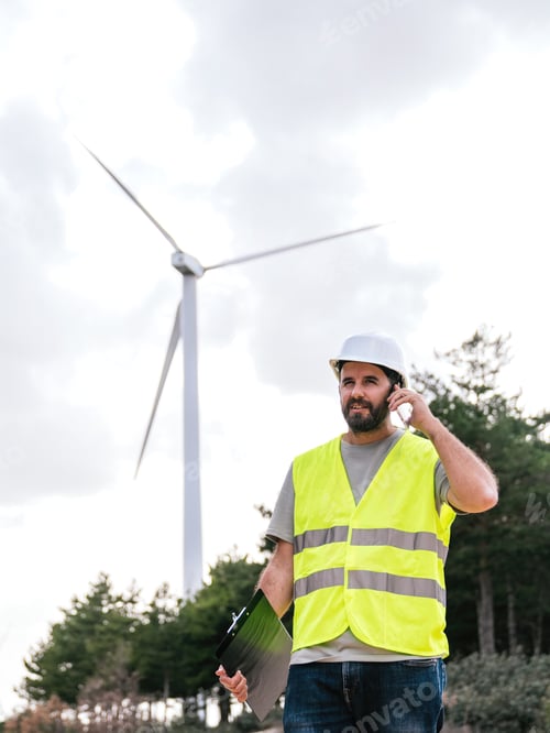 Preview: Wind turbine technician talking on the phone, holding clipboard, wearing safety vest and helmet.