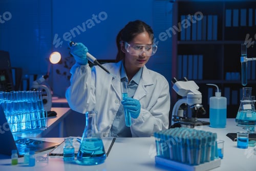 Preview: Female scientist using a micropipette to transfer blue liquid into a test tube, working late in a mo