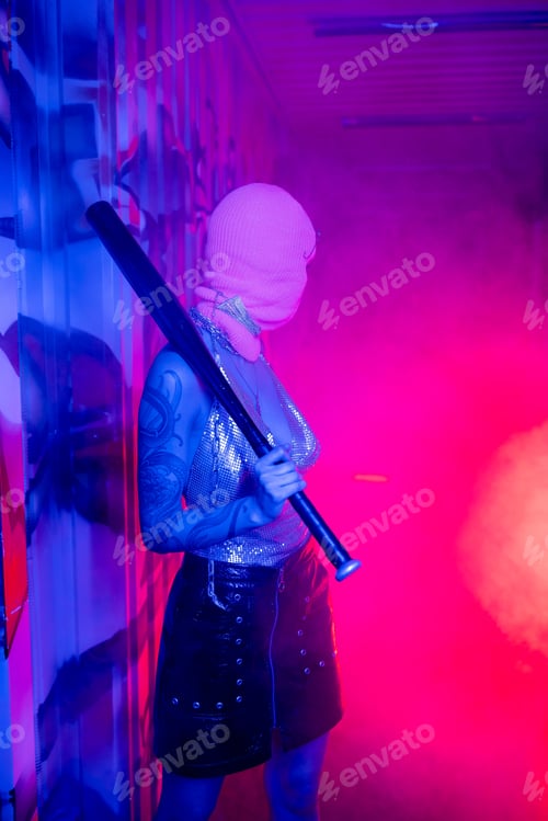 Preview: sexy woman in balaclava standing with baseball bat in purple light and looking at pink smoke