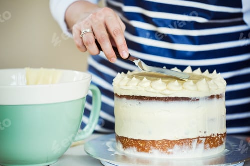 Preview: Close up of person wearing a blue and white stripy apron spreading cream over the top of a cake.