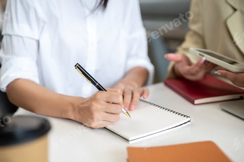 Preview: Close up of a woman student's hands holding pen writing in notebook aside teacher on table.