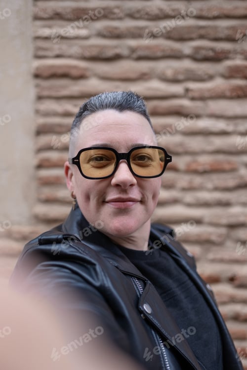 Preview: Smiling woman with yellow sunglasses taking a selfie in front of brick wall