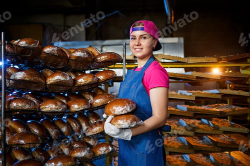 Preview: Smiling baker girl in apron holding bread and looking at camera. Young woman is a chef holding