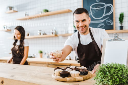 Preview: selective focus of handsome barista giving cupcake wile cashier working in coffee house