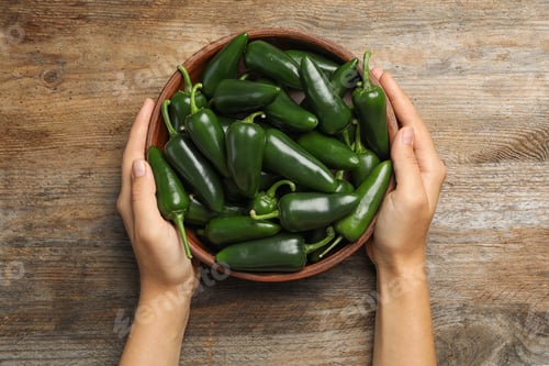 Preview: Young woman holding bowl with green hot chili peppers at wooden table, top view