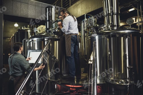 Preview: Male brewers in aprons working with tanks and industrial equipment at the brewery