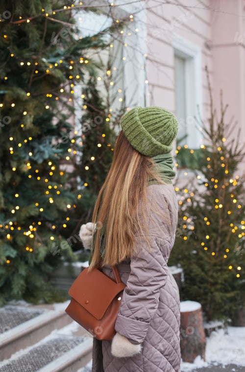 Preview: Stylish woman in winter standing near store or cafe decorated with green christmas trees and lights