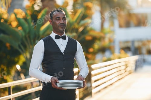 Preview: Holds empty plate. Black waiter in formal clothes is at his work outdoors at sunny daytime