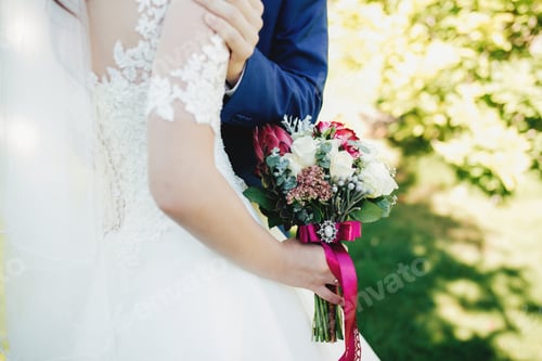 Preview: Bride in white dress holds wedding bouquet. Groom hugs bride. Blurred background