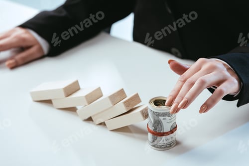 Preview: partial view of businesswoman sitting at table with fallen row of wooden blocks and money roll